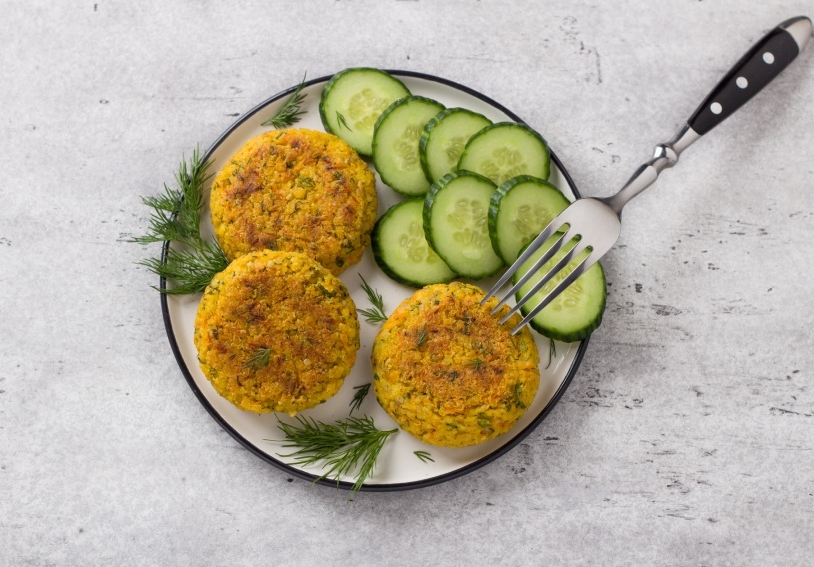 Delicious millet cutlets with carrots and seeds served with cucumber and herbs on a gray textured background, top view. Homemade vegan food